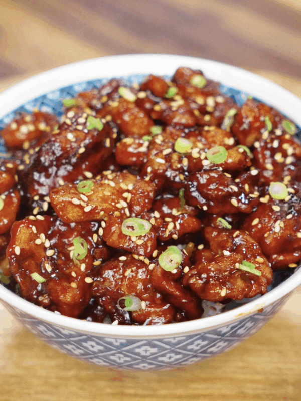 A full bowl of crispy sesame chicken coated in a thick, shiny glaze and topped with scallions and sesame seeds.