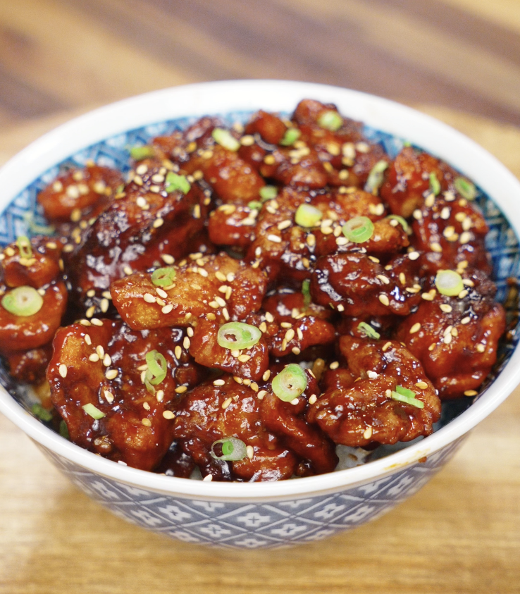 A full bowl of crispy sesame chicken coated in a thick, shiny glaze and topped with scallions and sesame seeds.