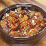 A bowl of sticky sweet pork belly bites glazed in a glossy honey-soy sauce, served over steamed rice and topped with sliced scallions.