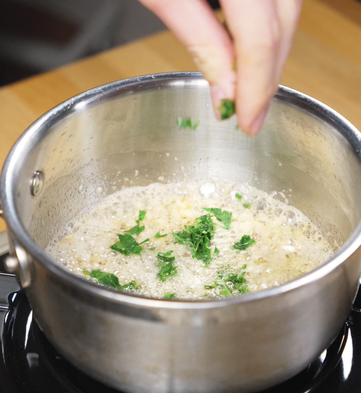 Chopped parsley is being added to a bubbling garlic butter sauce in a stainless steel saucepan.