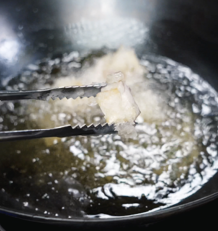 A single battered chicken piece is lifted from hot oil during the first fry at 350°F, still pale and crisping up.