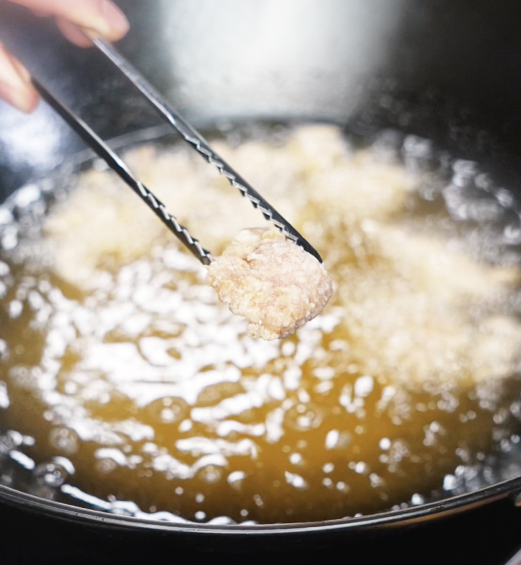 A piece of lightly fried chicken is lifted from hot oil, showing the pale first-fry texture before crisping.