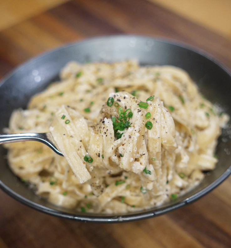 A fork lifts a rich, creamy bite of garlic fettuccini Alfredo, topped with herbs and black pepper.
