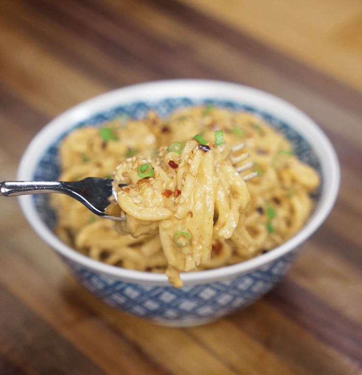 A fork lifts a glossy swirl of creamy peanut butter noodles from the bowl, coated in sauce and topped with scallions and sesame seeds.