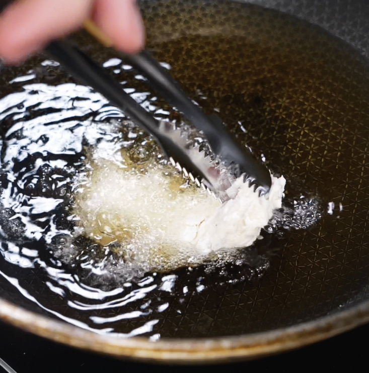 A pair of tongs gently lowers a battered chicken tender into hot oil, starting the deep-frying process until golden and crispy.