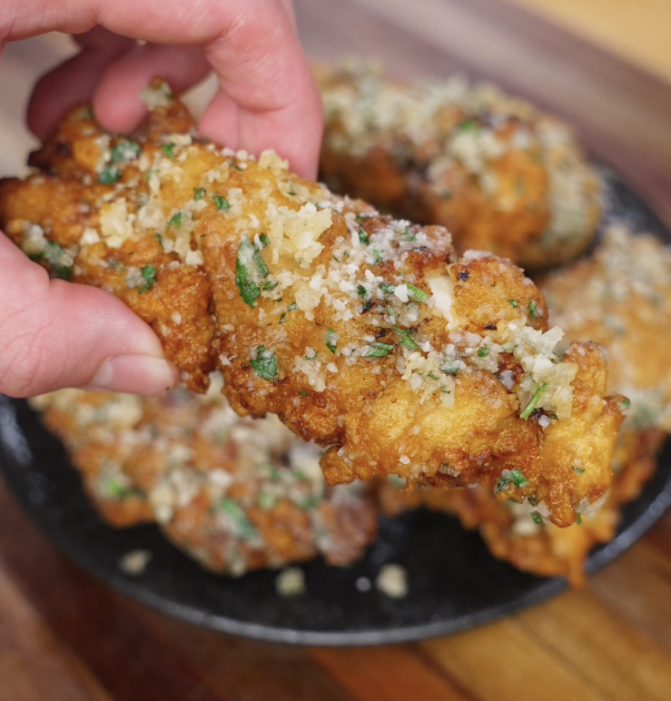 A single garlic parmesan chicken tender is held up to the camera, showing its crispy coating glistening with garlic butter and cheese.