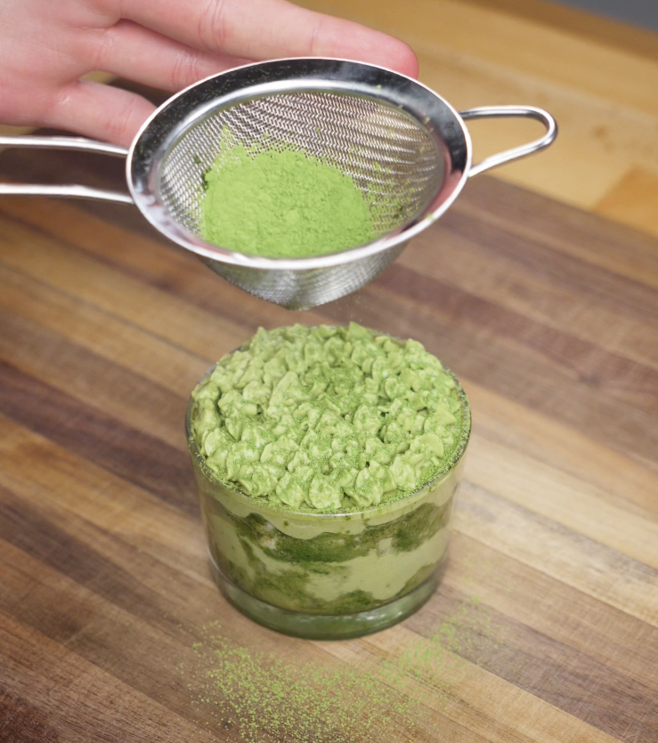 Three individual matcha tiramisu cups are topped with piped matcha cream and a dusting of extra matcha powder.