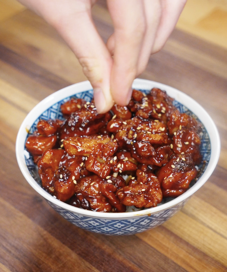 A hand sprinkles sesame seeds over a finished bowl of saucy sesame chicken.