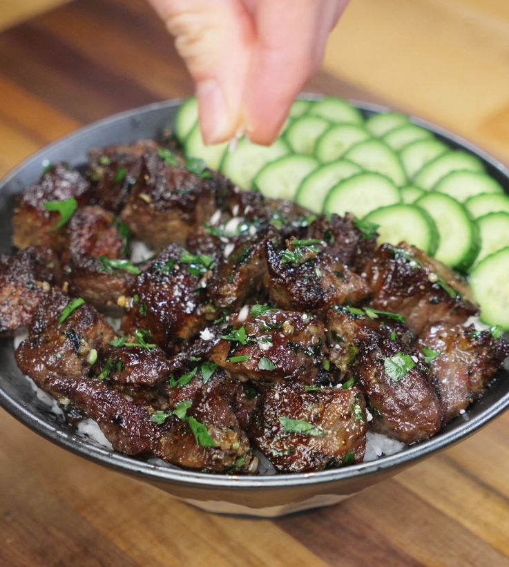 A hand sprinkles parmesan cheese over the finished garlic steak bites rice bowl, completing the dish.