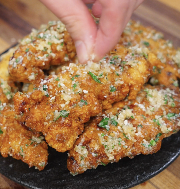A hand sprinkles extra parmesan over a plate of garlic parmesan chicken tenders, finishing them with cheesy, herby flair.