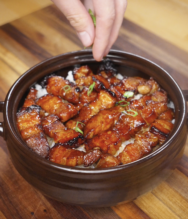 A hand sprinkles thinly sliced scallions over the finished bowl of sticky sweet pork belly for a fresh finish.