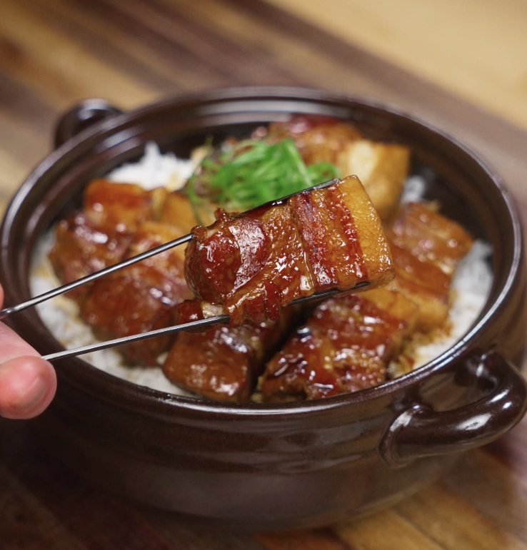 A piece of juicy pork belly is lifted with chopsticks, showing the rich caramelized sauce and tender texture.