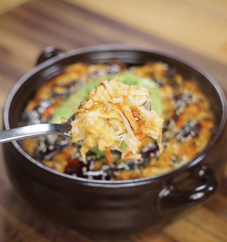 A spoon lifts a creamy, golden bite of sushi bake filled with crab, rice, and sauce from the baking dish.