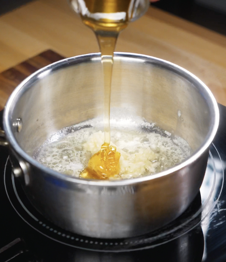 Golden honey is poured into a saucepan of melted butter and sautéed garlic.