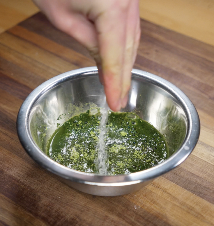 Matcha powder and sugar are sprinkled into a bowl of hot water to prepare the soaking liquid for ladyfingers.