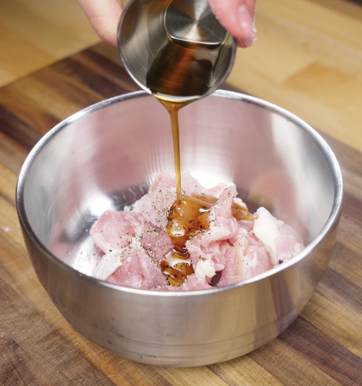 Soy sauce and seasonings are poured over raw chicken thigh chunks in a metal mixing bowl to marinate before frying.