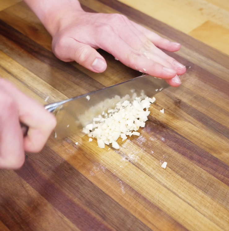 A close-up of hands finely mincing fresh garlic cloves on a wooden cutting board with a sharp knife.
