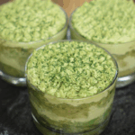 Three glass cups filled with layered matcha tiramisu, topped with piped matcha cream and a dusting of matcha powder, arranged on a dark countertop for a striking contrast.