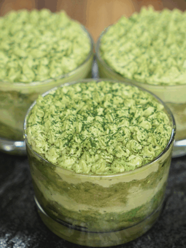 Three glass cups filled with layered matcha tiramisu, topped with piped matcha cream and a dusting of matcha powder, arranged on a dark countertop for a striking contrast.