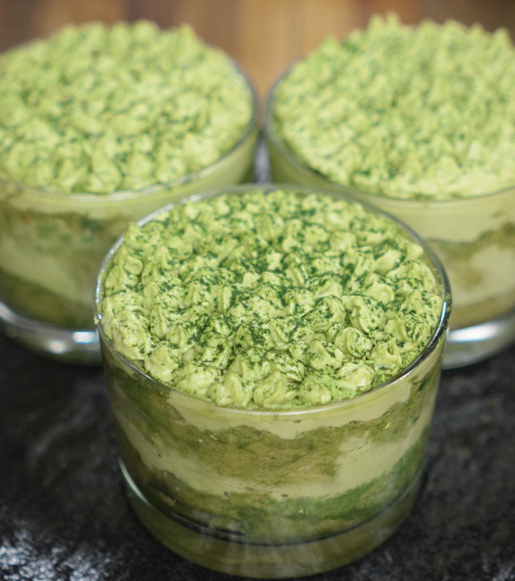 Three glass cups filled with layered matcha tiramisu, topped with piped matcha cream and a dusting of matcha powder, arranged on a dark countertop for a striking contrast.