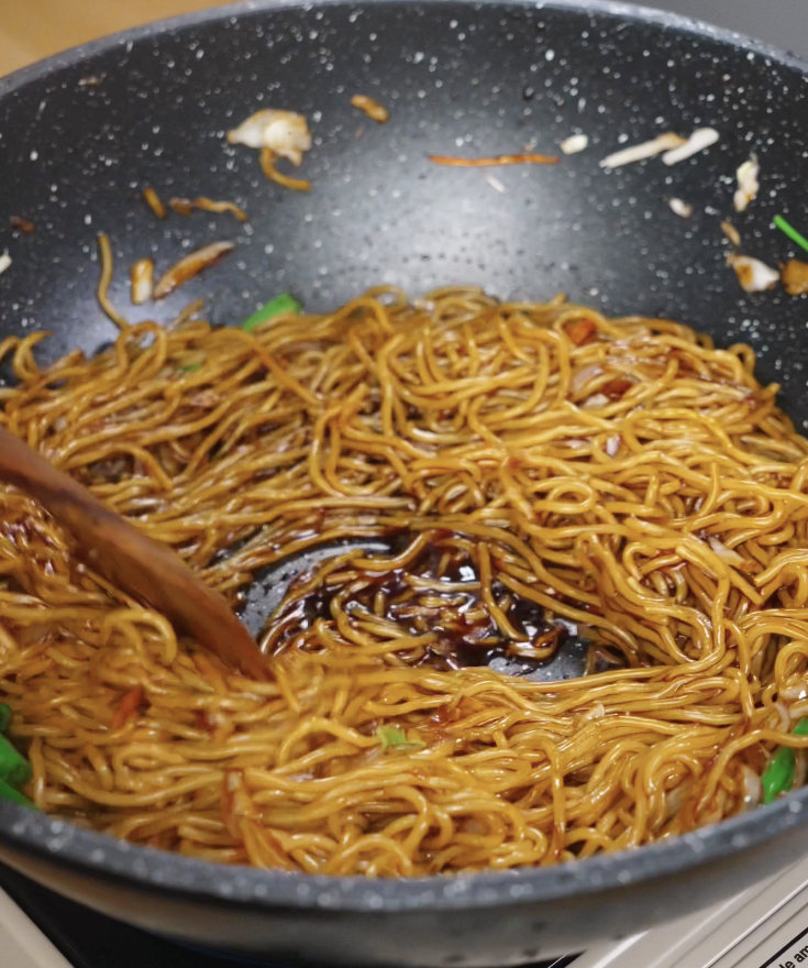 Chow mein noodles are tossed in a wok with sauce and vegetables, forming a ring as the sauce coats the bottom.