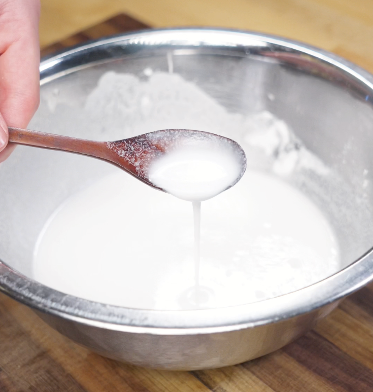A spoon drips a slightly thin white batter made from rice flour, potato starch, salt, and water into a large bowl.