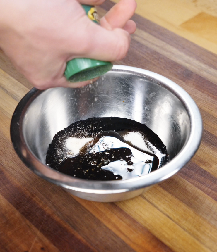 A hand adds white pepper from a green spice jar into a bowl of soy sauce, sugar, sesame oil, and shaoxing wine to make chow mein sauce.