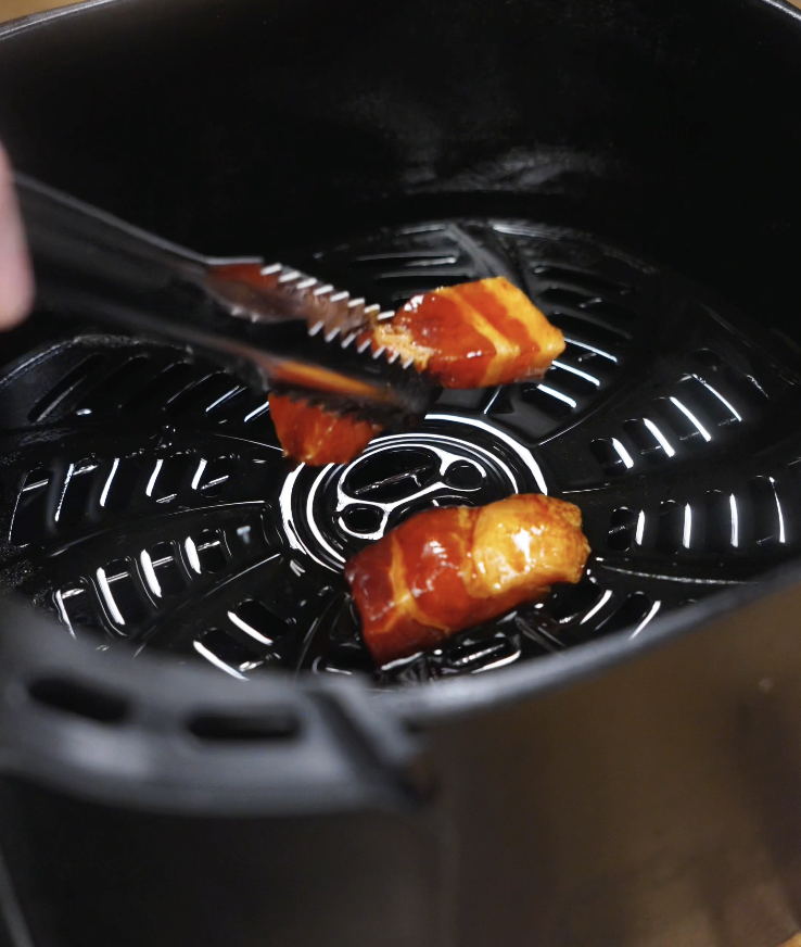 Tongs lowering marinated pork belly pieces into the air fryer basket lined with parchment, ready to cook.