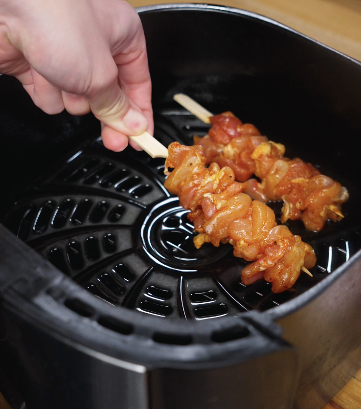 Chicken skewers are carefully laid inside an oiled air fryer basket before cooking.
