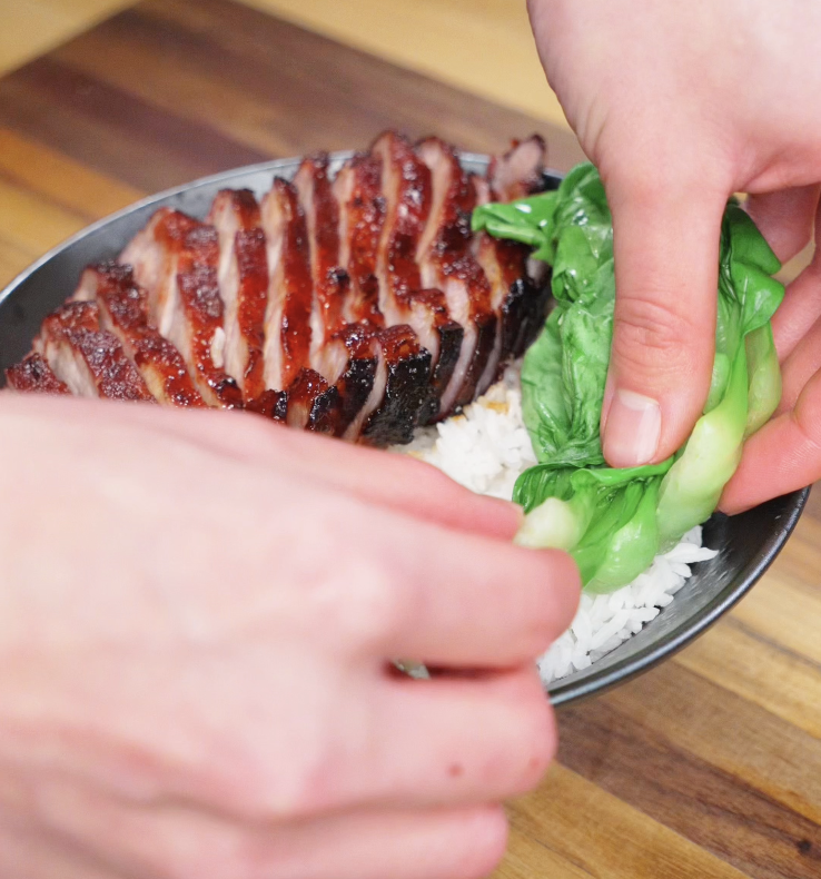 Hands placing steamed leafy greens beside char siu slices and rice in a serving bowl.