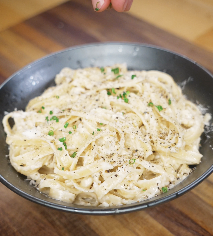 The saucy garlic fettuccini is plated in a wide bowl with visible chives and cracked pepper on top.