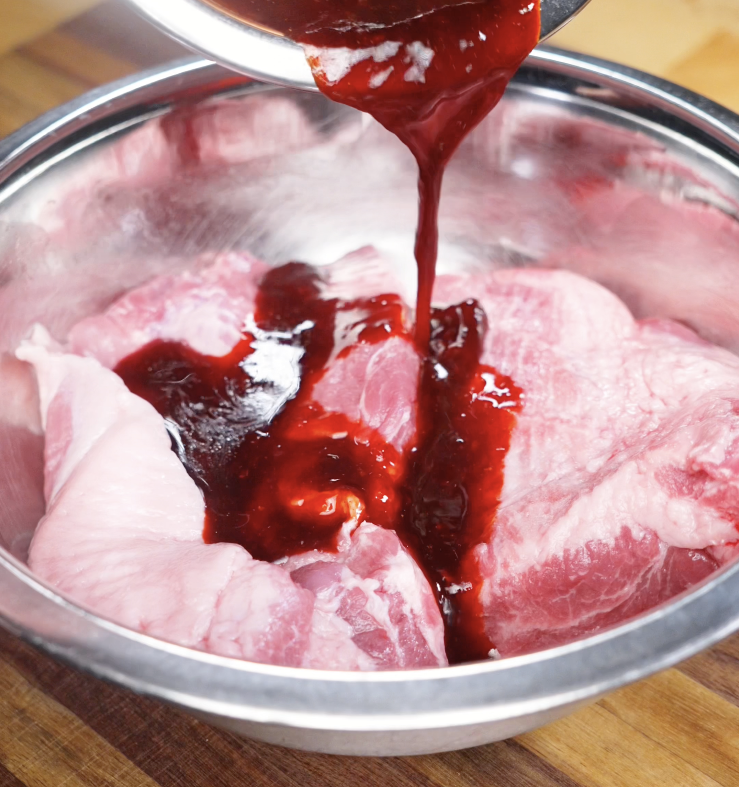 A rich red char siu marinade being poured over raw pork shoulder strips in a mixing bowl.