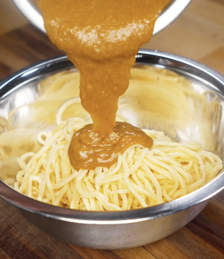 A thick, creamy peanut sauce is poured over a bowl of freshly cooked noodles, ready to be mixed.