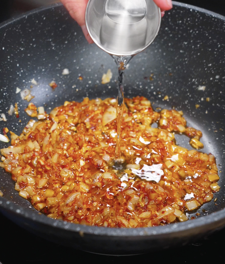 A hand pours clear vodka into the sautéed onion and chili paste mixture to deglaze and add depth.