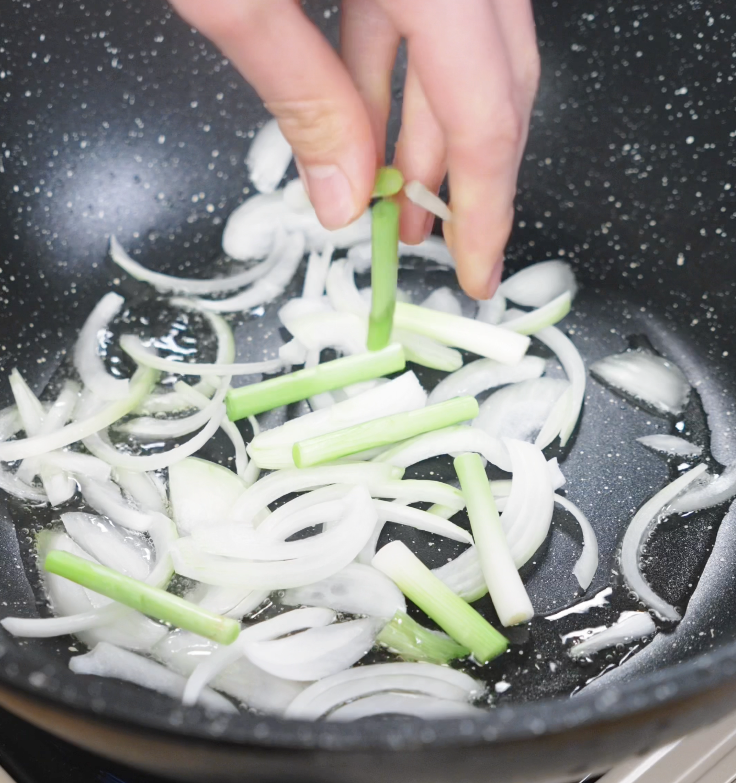 Sliced white onions and scallion whites are being sautéed in a hot oiled wok as the base for chow mein.