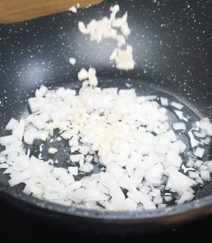 Diced white onions and minced garlic are sizzling in oil inside a nonstick pan to build the base for the sauce.