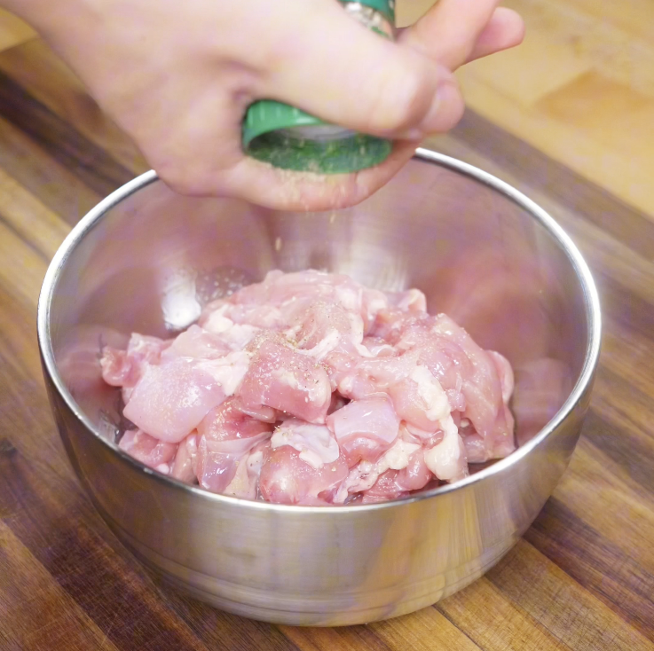 Salt and white pepper being sprinkled onto raw chicken chunks during the marinating step.