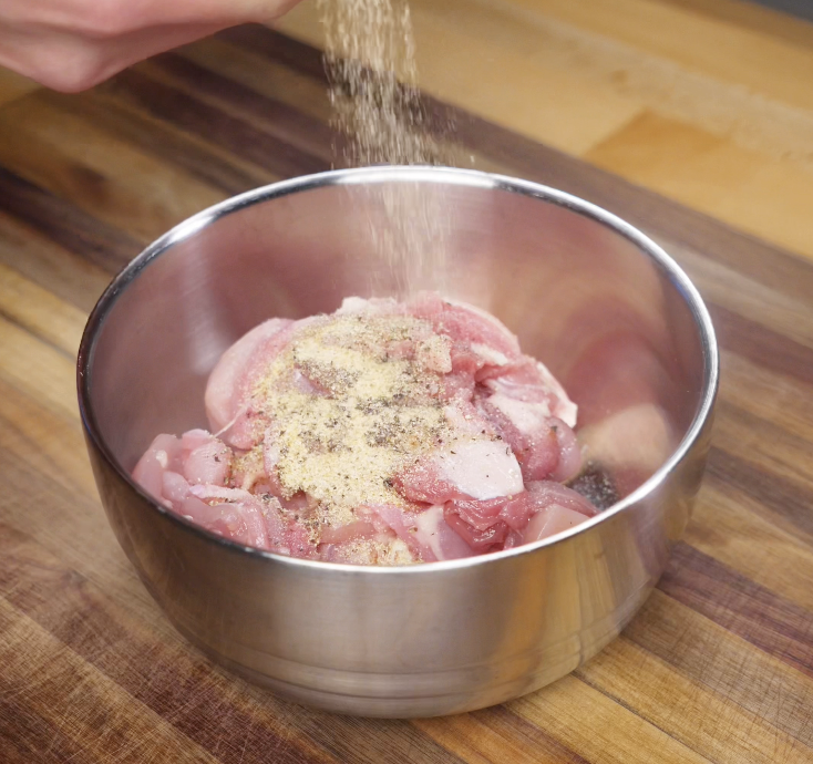 Garlic powder, paprika, salt, and pepper are sprinkled over raw chicken pieces in a metal bowl.