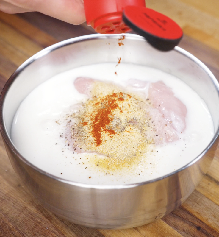 Paprika and garlic powder are sprinkled into a bowl of buttermilk and raw chicken tenders, creating the flavorful marinade base.