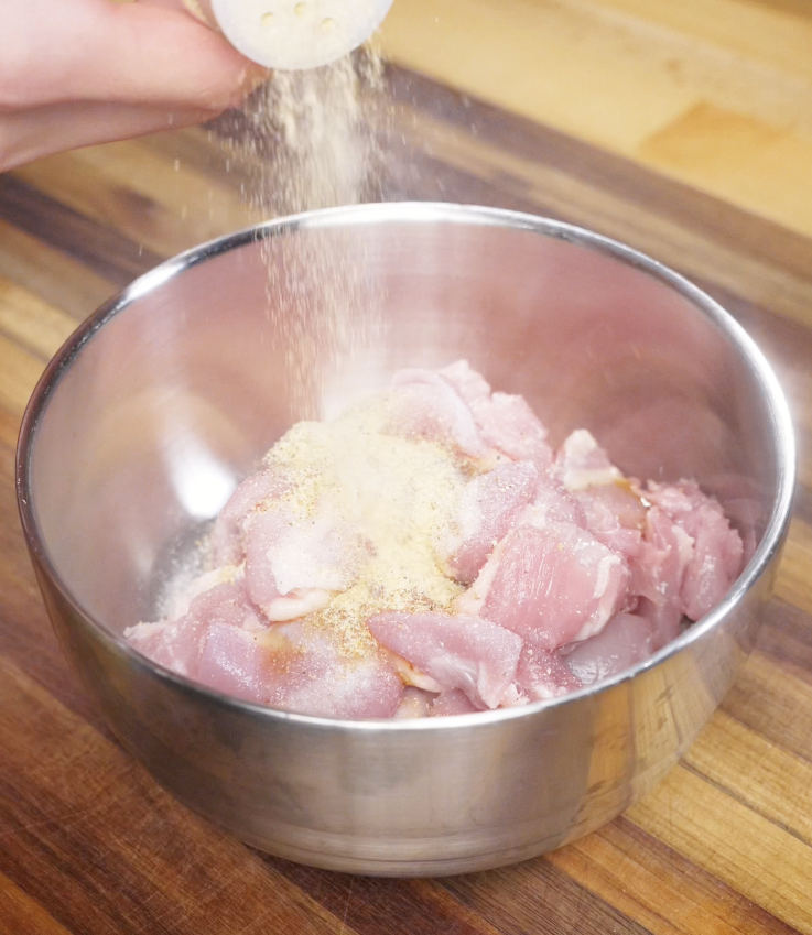 Raw chicken thigh pieces in a metal bowl being sprinkled with salt, white pepper, garlic powder, and sesame oil.