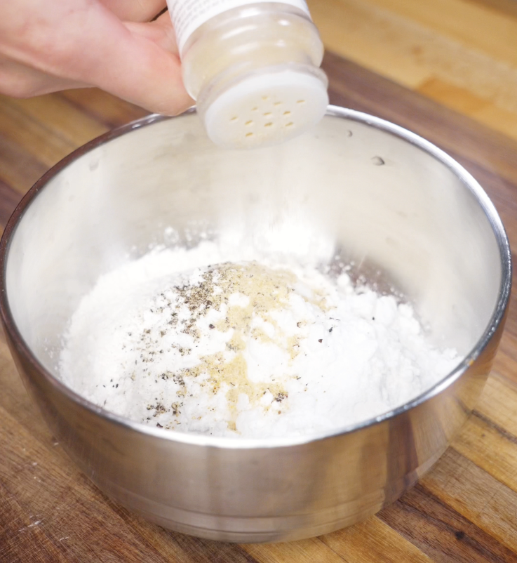 Garlic powder is shaken into a bowl of flour, potato starch, salt, and pepper to season the dry dredge for crispy chicken tenders.