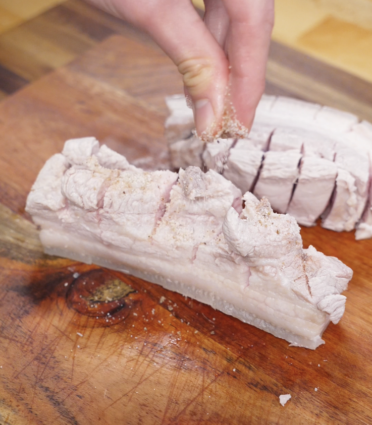 A hand sprinkles spice rub onto the meat side of partially cooked pork belly on a wooden cutting board.