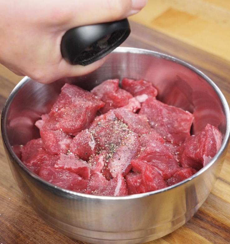 Freshly ground black pepper is sprinkled over cubed steak in a metal bowl for marination.