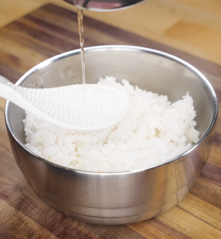 Warm sushi rice is mixed with vinegar seasoning using a rice paddle in a stainless steel bowl.