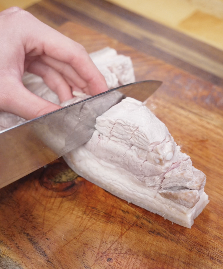 A hand slices through a block of boiled pork belly on a wooden cutting board, showing the tender layers of meat and fat.