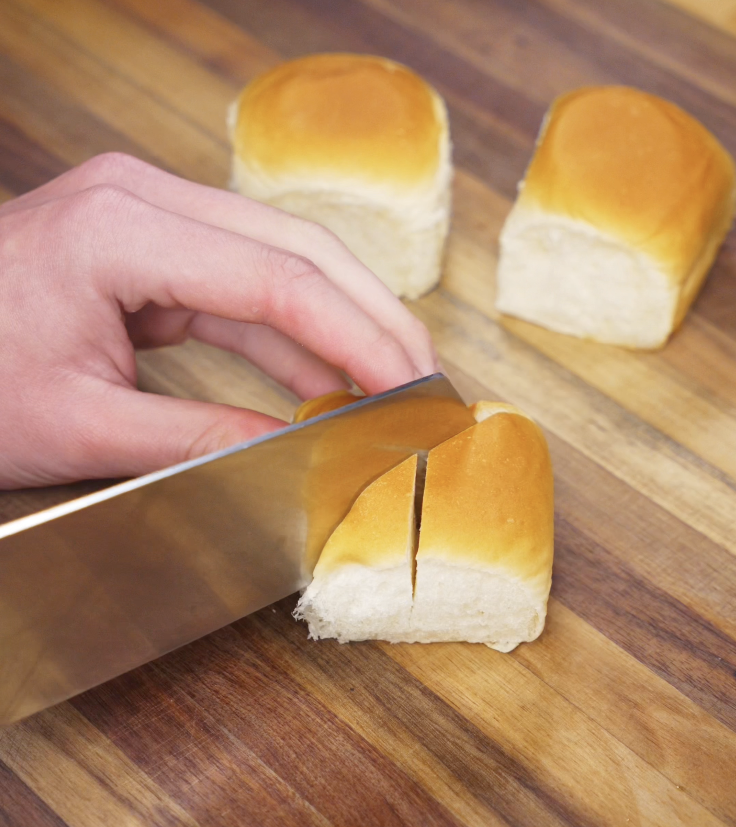 A hand carefully cuts deep slits into a soft bread roll on a wooden cutting board, prepping it for filling.