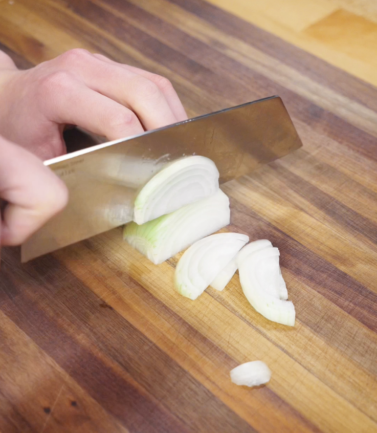 A hand slices a white onion into thin strips on a wooden cutting board using a large chef’s knife.