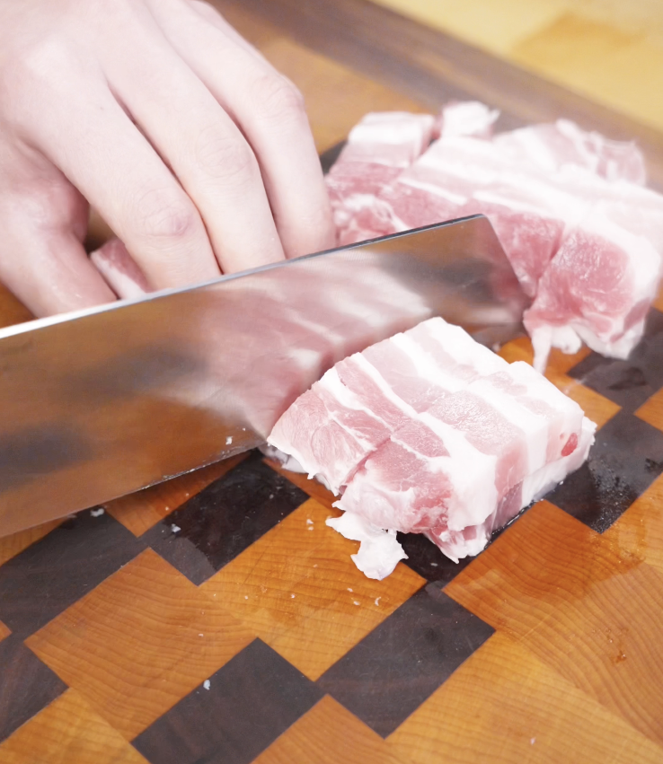 A hand slices raw pork belly into thick, even chunks on a checkered wooden cutting board in preparation for marinating.