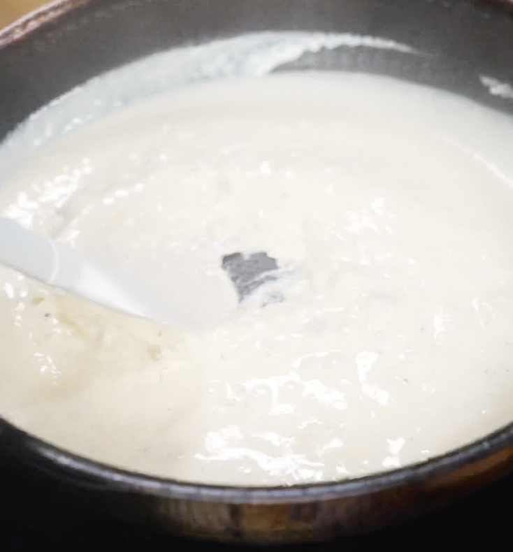 The Alfredo sauce is stirred with a spatula as it thickens into a smooth, creamy texture before adding pasta.