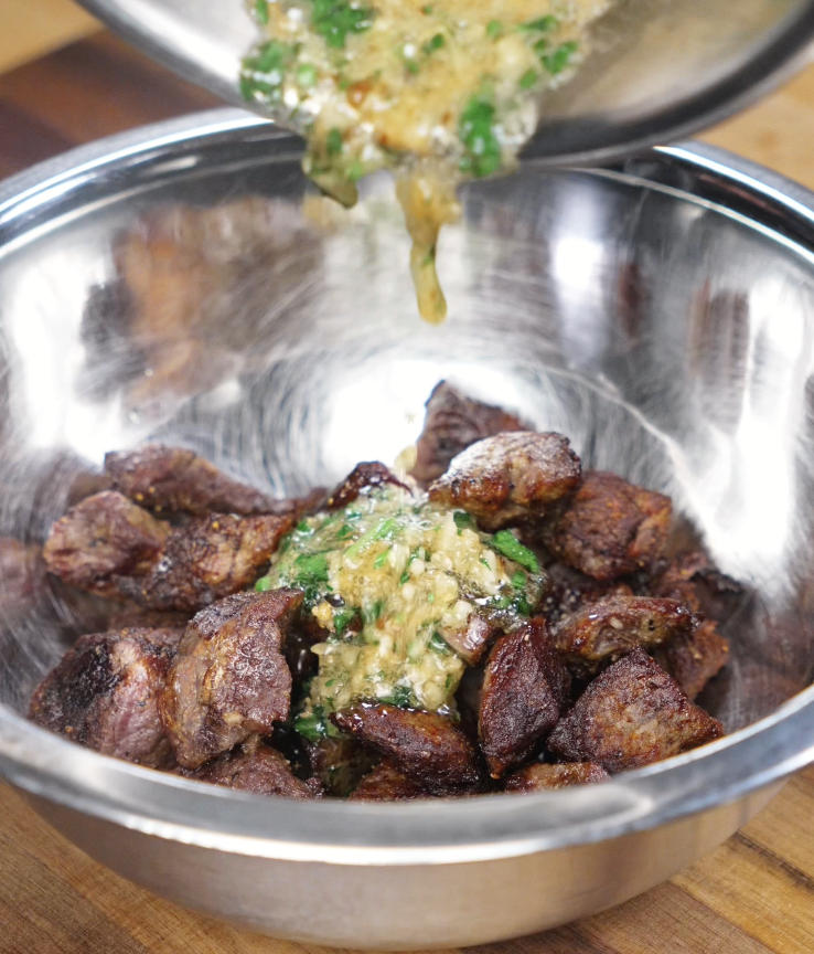 Hot garlic butter is being poured over crispy steak bites in a metal mixing bowl for a final toss.